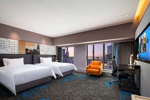 Ein Hotelzimmer mit zwei Betten, einem Schreibtisch, einem Stuhl und einem großen Fenster mit Blick auf die Stadt.