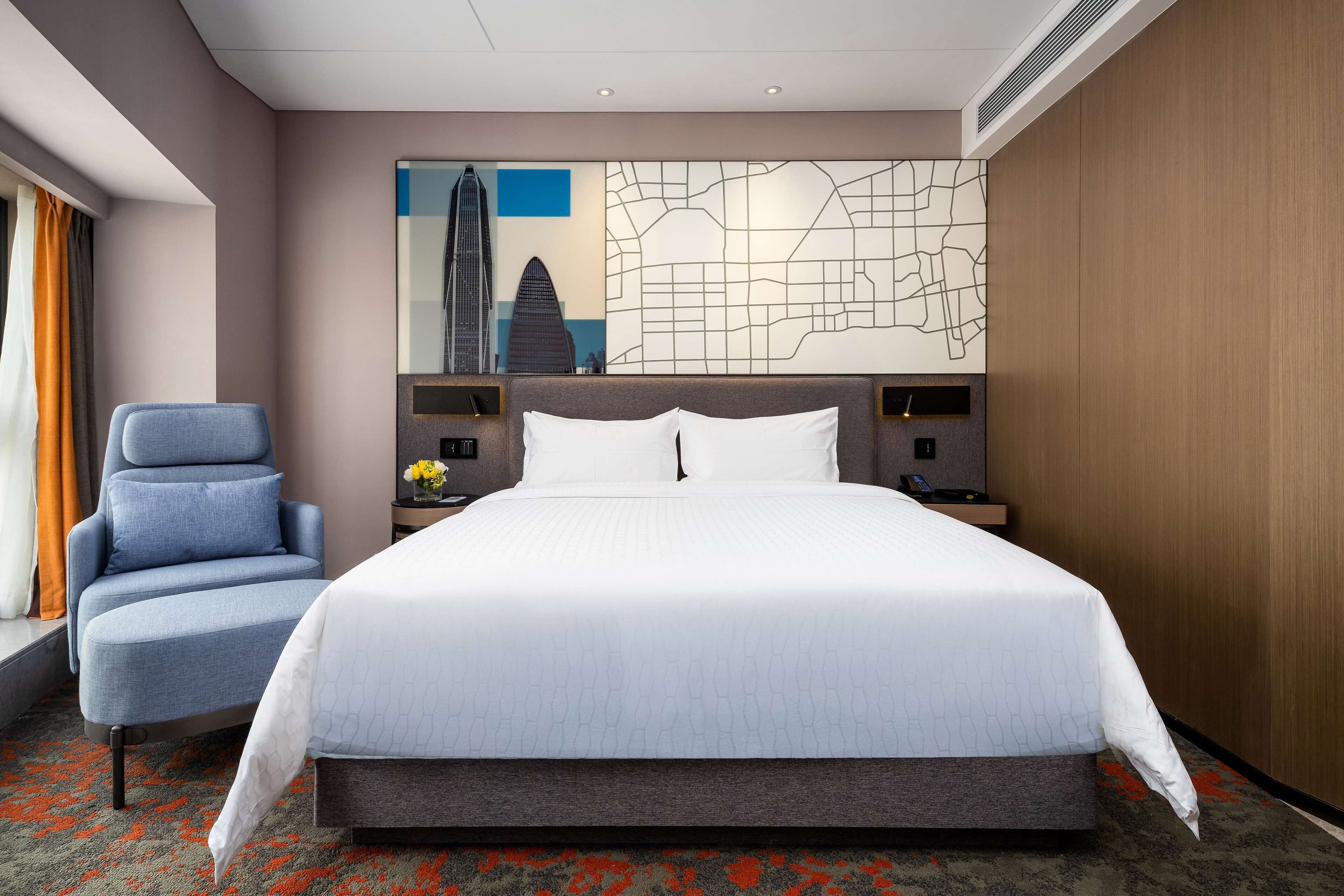 A modern hotel room with a large bed, a blue armchair, a map artwork on the wall, and a cityscape view.