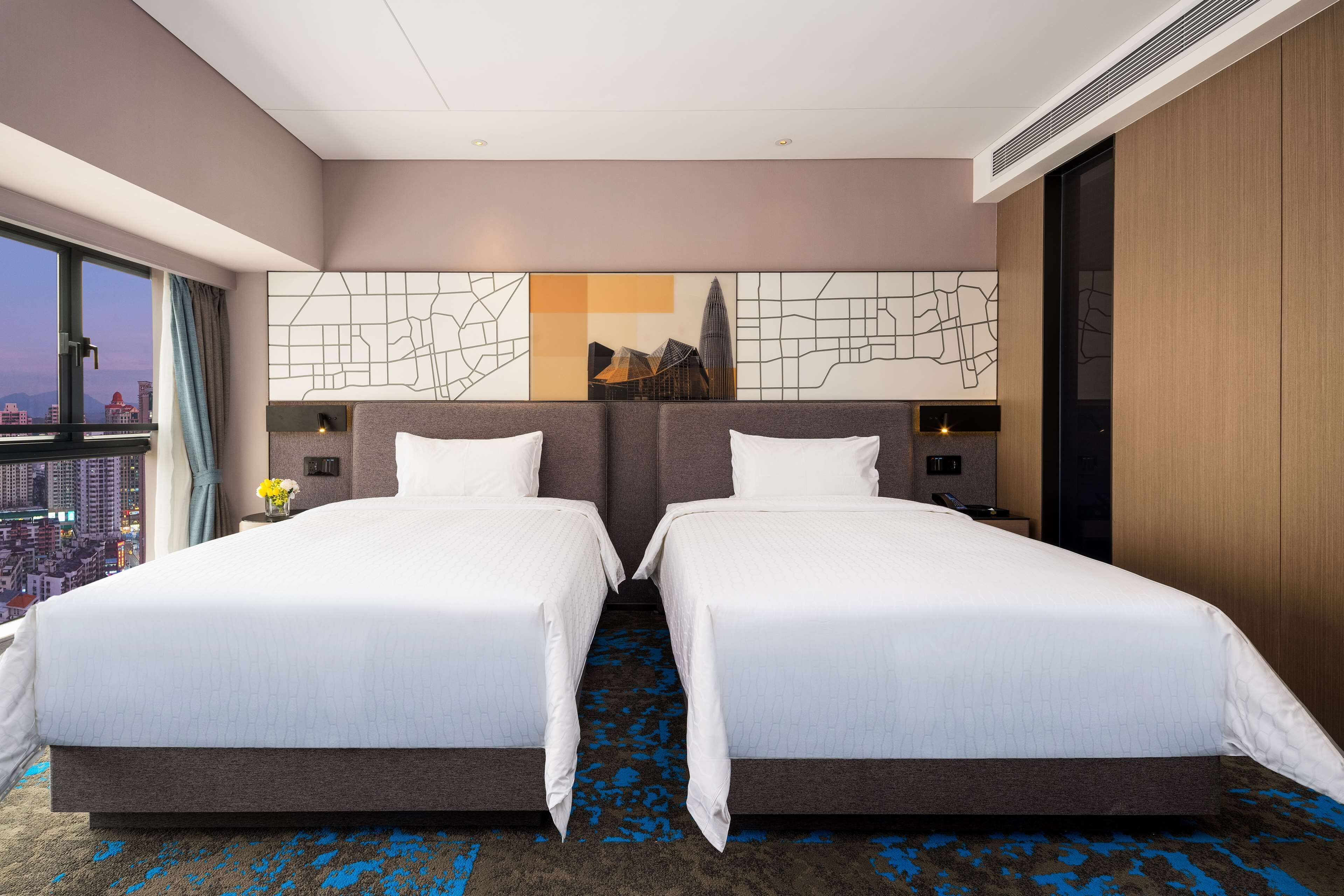 Une chambre d’hôtel avec deux lits, une vue sur la ville, une grande œuvre d’art abstrait accrochée au mur et une moquette à motifs bleus.