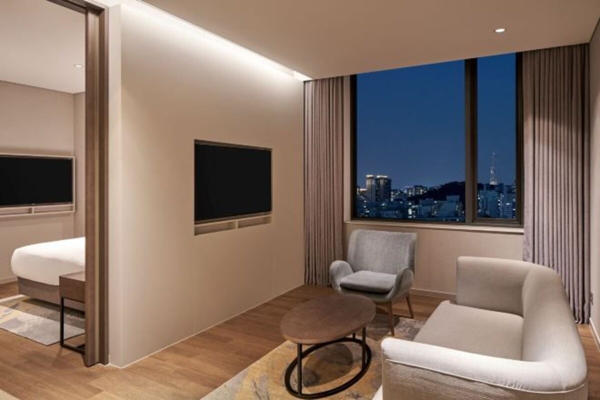 A modern hotel room with a city view, a flat-screen TV, a sofa, a chair, and a small table.