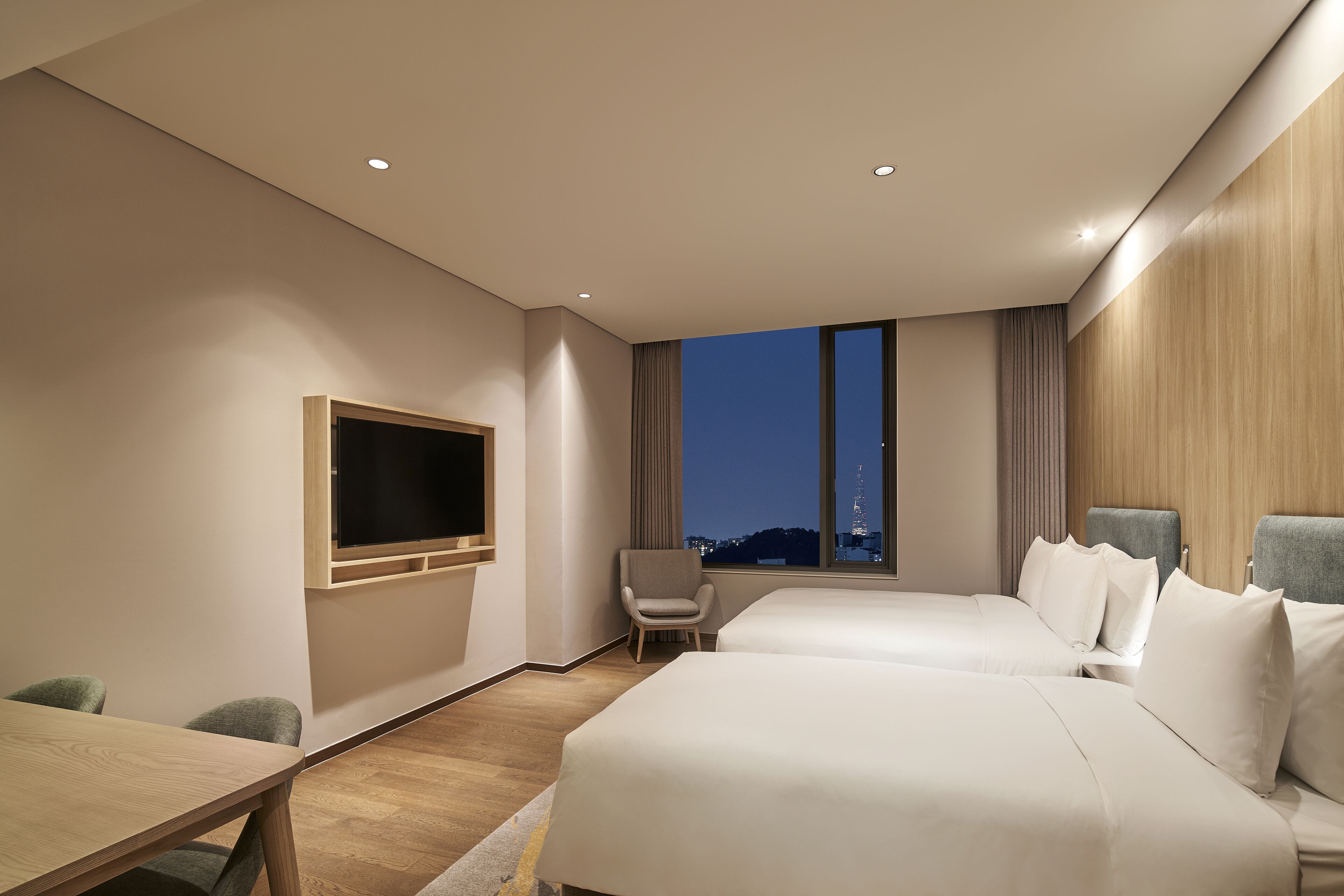 A modern hotel room with two beds, a TV mounted on the wall, a desk, and a view of a cityscape at night.