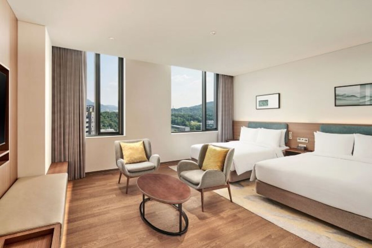 A hotel room with a large bed, two armchairs, a small round table, and a view of the outdoors through large windows.