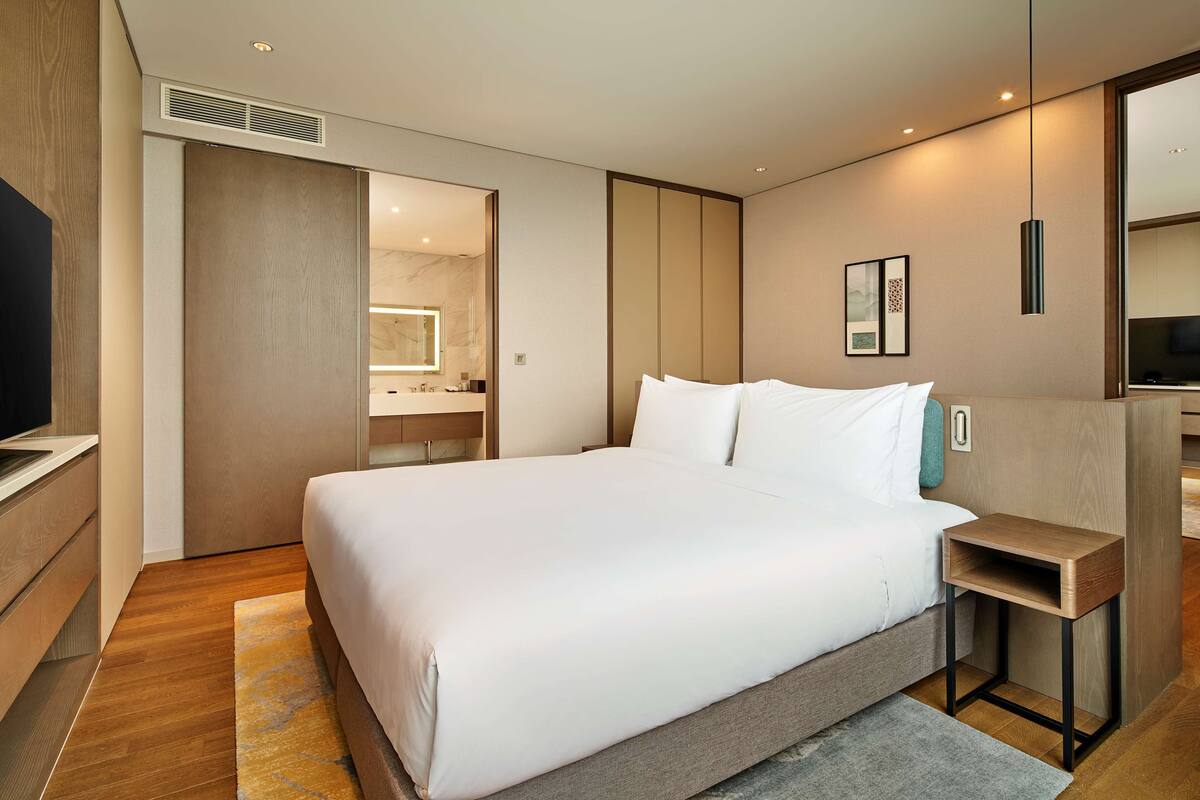Une chambre d’hôtel moderne dotée d’un grand lit, d’une table de chevet, d’un téléviseur intégré et d’une salle de bain visible à travers une porte ouverte.