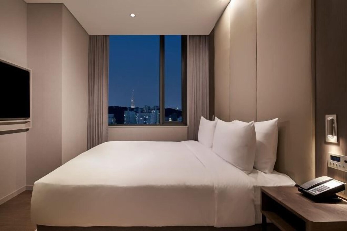 Une chambre d’hôtel avec un grand lit, un téléphone sur la table de chevet et une vue sur le paysage urbain illuminé la nuit.