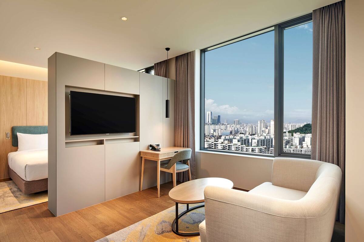 Une chambre d’hôtel moderne équipée d’un lit, d’un bureau, d’une chaise, d’une télévision et d’une grande fenêtre donnant sur la ville.