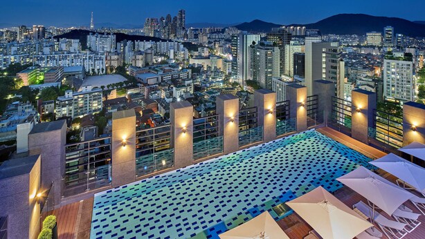 Ein Dachpool mit einem geometrischen Fliesendesign, umgeben von modernen Gebäuden und einer Stadtlandschaft im Hintergrund.