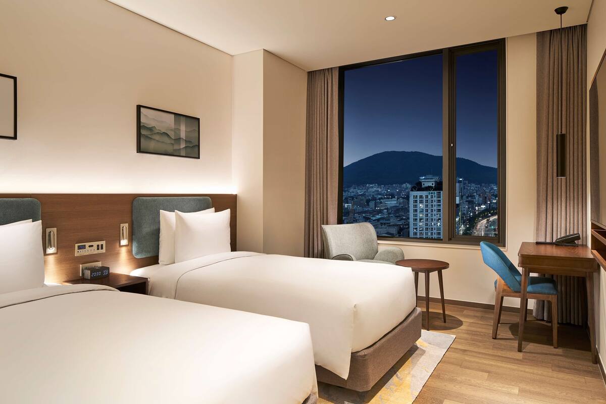 Une chambre d’hôtel avec deux lits, un bureau, une chaise et une grande fenêtre donnant sur le paysage urbain.