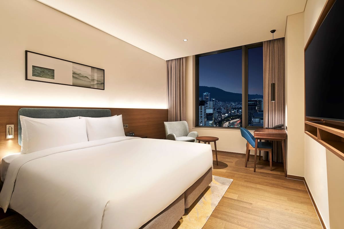 Une chambre d’hôtel avec un grand lit, un bureau, une chaise et une vue sur le paysage urbain illuminé la nuit.