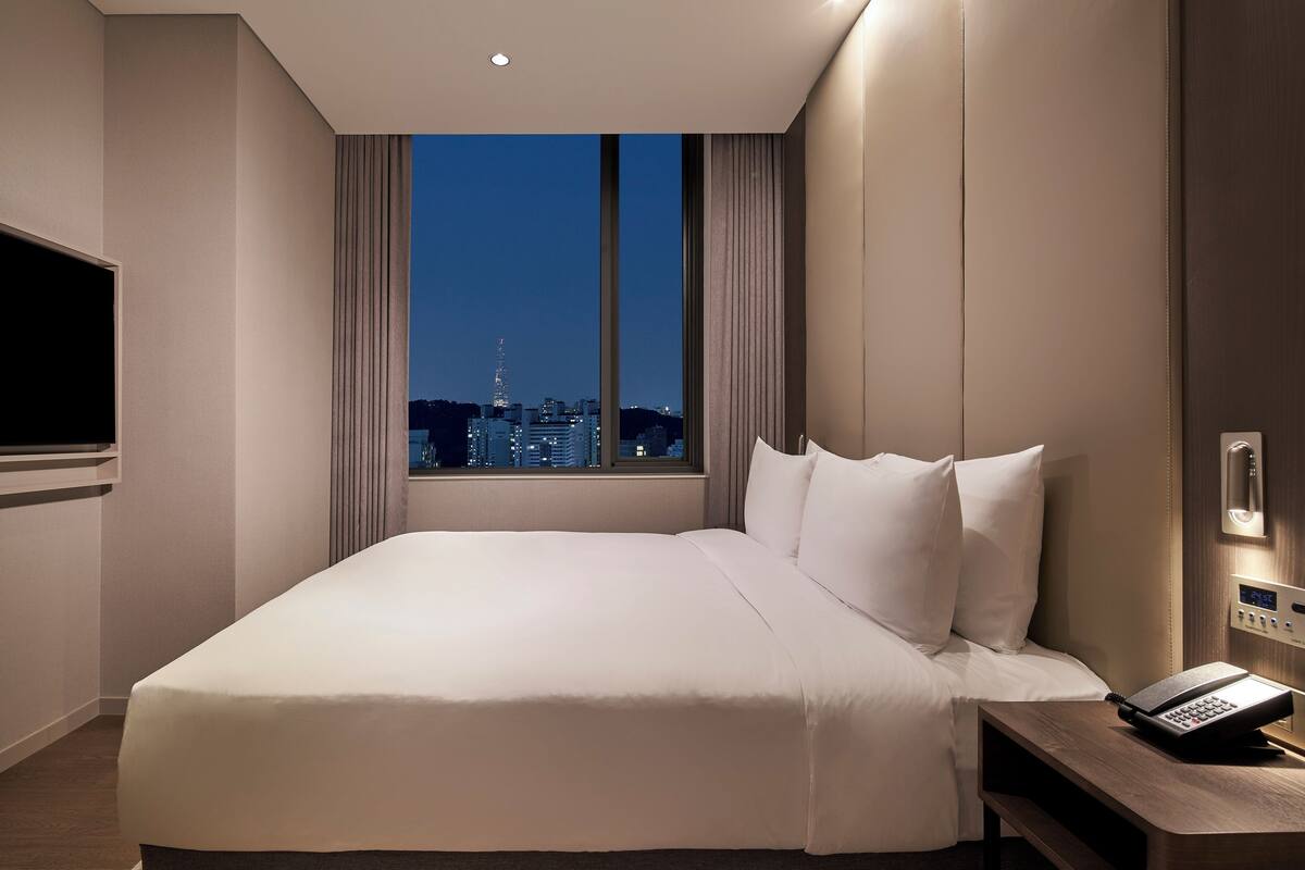 Une chambre d’hôtel avec un grand lit, un téléphone sur la table de chevet et une vue sur le paysage urbain par la fenêtre.