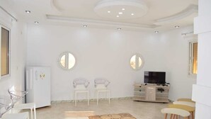 A spacious living room with a white color scheme, a flat-screen TV, two chairs, a small table, and a refrigerator. - Tunisia, royal duplex (Salakta)