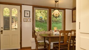 A dining area with a wooden table and chairs, a chandelier, and a view of greenery through large windows. - Charming, historic, Creekside Cabin nestled at the base of Pikes Peak in Cascade (Cascade,)