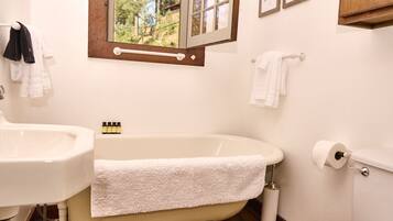 A bathroom with a clawfoot bathtub, a white sink, and a toilet. There are towels hanging on the wall and a window with a view of trees.