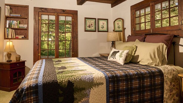 A bedroom with a large bed, a wooden headboard, a nightstand, a bookshelf, and a window with a view of trees. - Charming, historic, Creekside Cabin nestled at the base of Pikes Peak in Cascade (Cascade,)