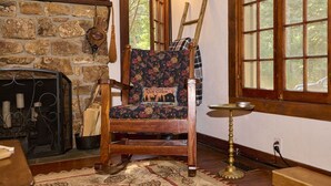A rustic cabin interior with a stone fireplace, a wooden chair with a floral cushion, and a window with a view of trees. - Charming, historic, Creekside Cabin nestled at the base of Pikes Peak in Cascade (Cascade,)