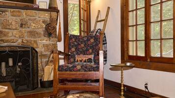 A rustic cabin interior with a stone fireplace, a wooden chair with a floral cushion, and a window with a view of trees.