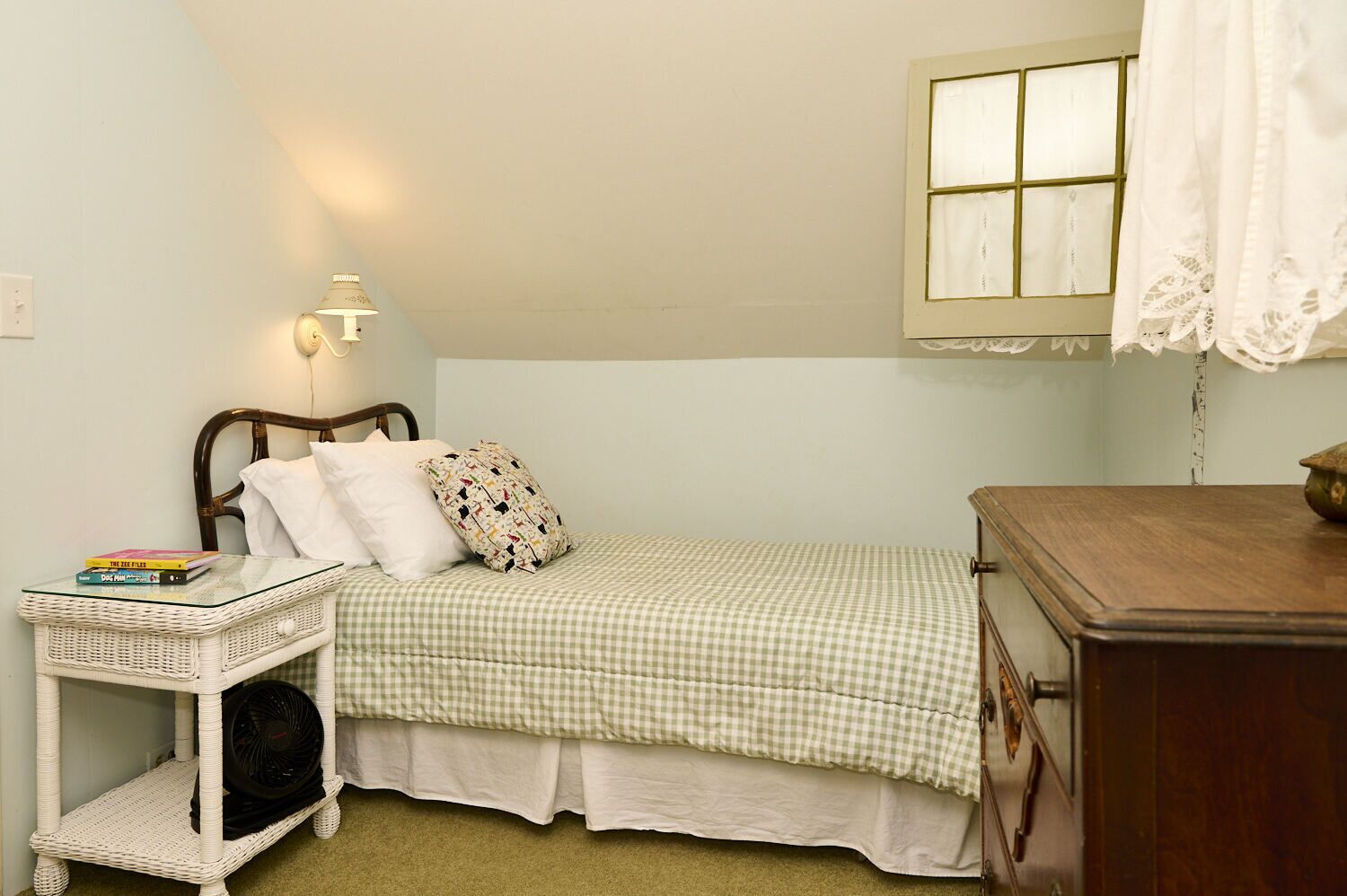 Um quarto pequeno e acolhedor com uma cama de solteiro, mesa de cabeceira com livros, abajur e janela com cortinas de renda.