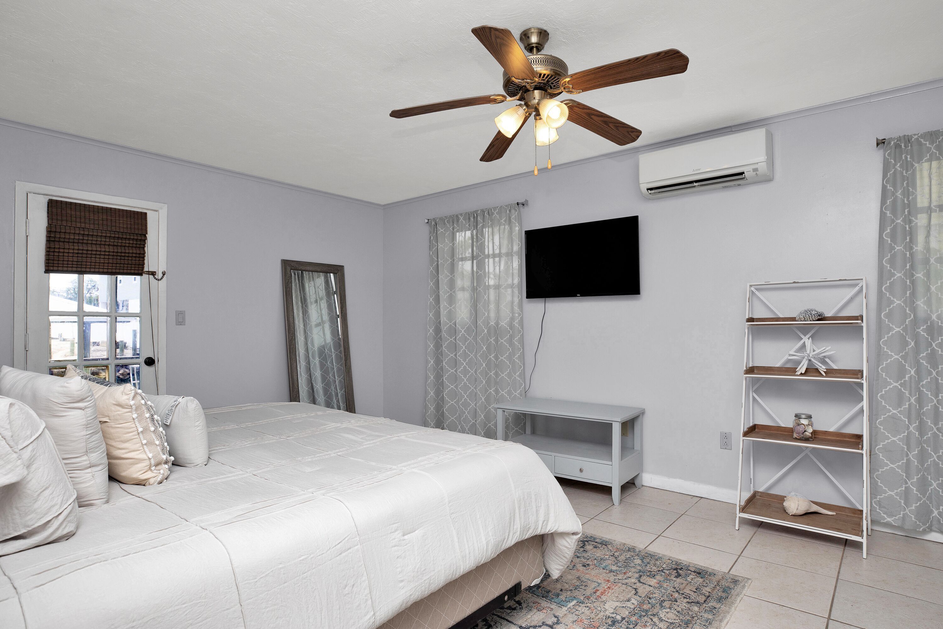 Une chambre à coucher avec un lit, un ventilateur de plafond, une télévision, une étagère et une fenêtre avec des rideaux.