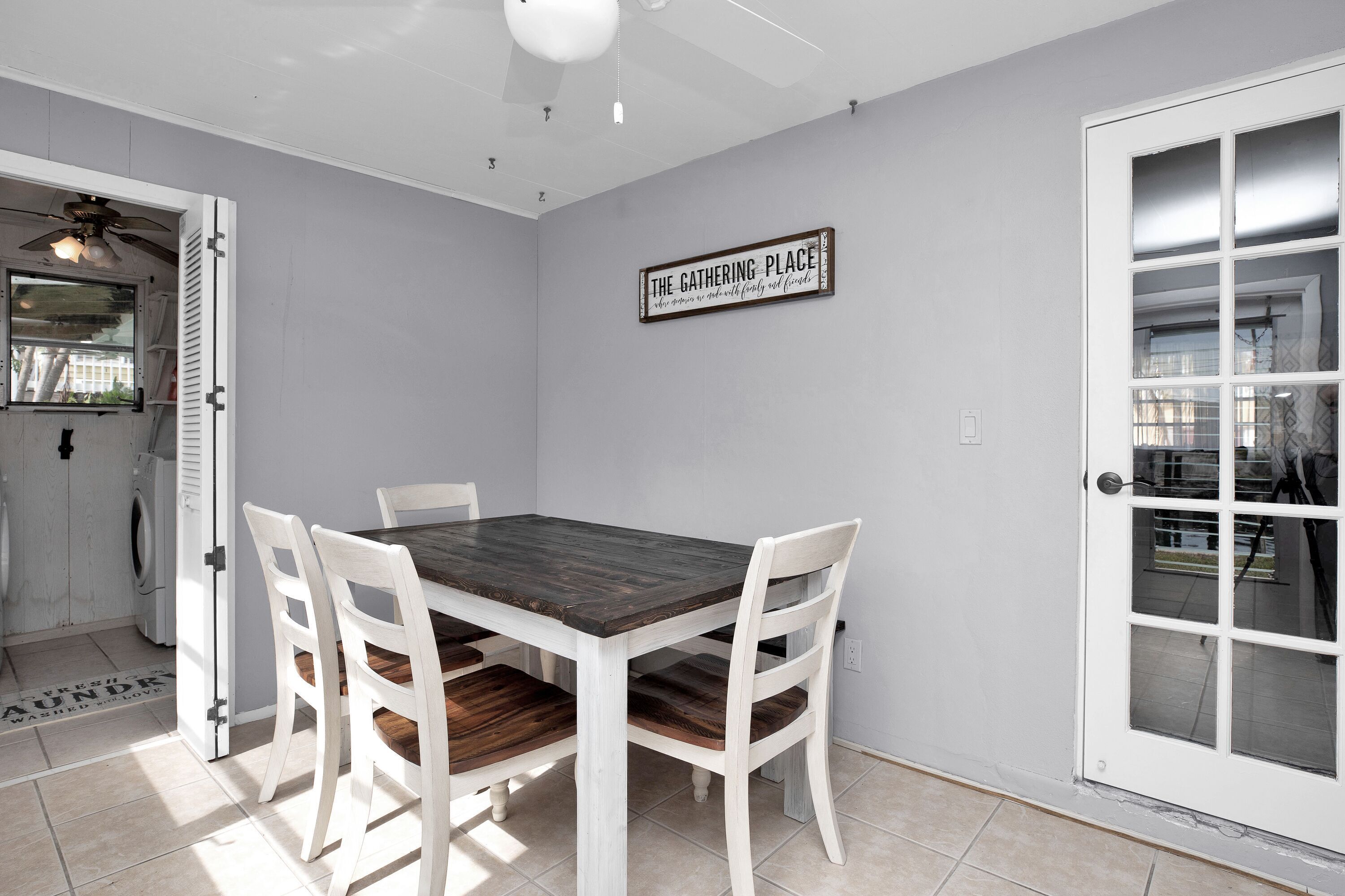 A dining area with a wooden table and white chairs, a ceiling fan, and a sign reading "THE GATHERING PLACE".