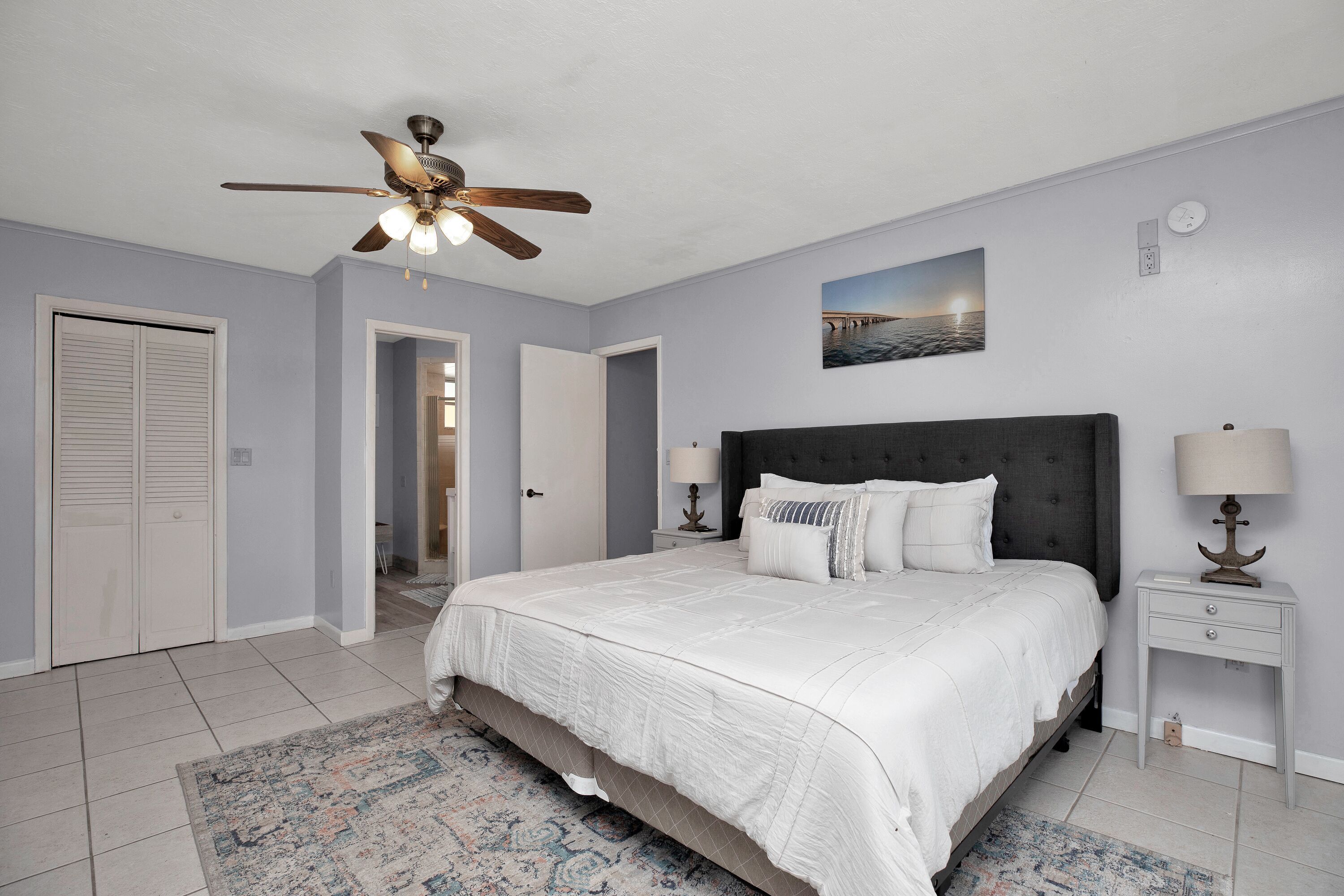 Une chambre à coucher avec un lit, un ventilateur de plafond, une table de chevet avec une lampe, une porte et une horloge murale.