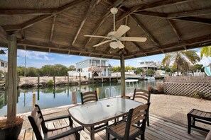 A covered outdoor seating area with a table and chairs, overlooking a body of water with boats and houses in the background. - Yellowtail Escape! 3/2 single family w. 60ft dock (Marathon)