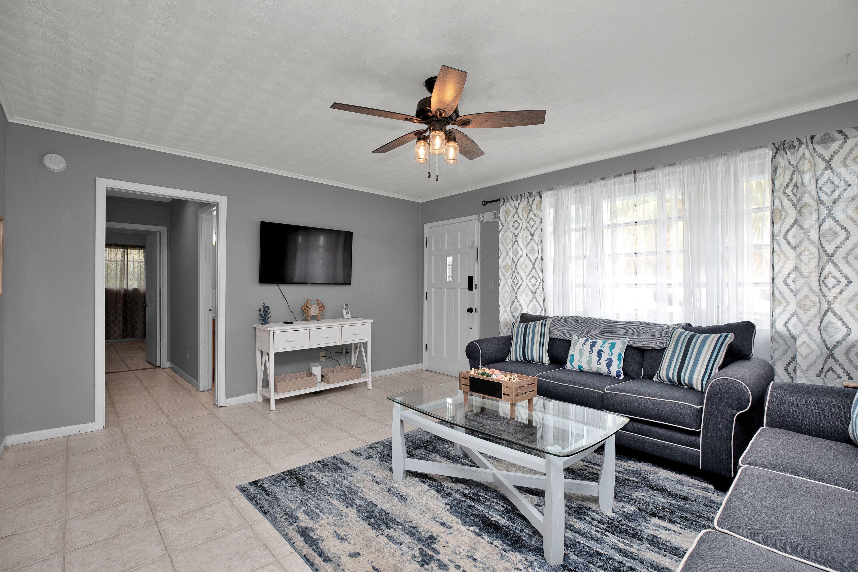 A modern living room with a grey sofa, a glass coffee table, a ceiling fan, and a patterned rug.