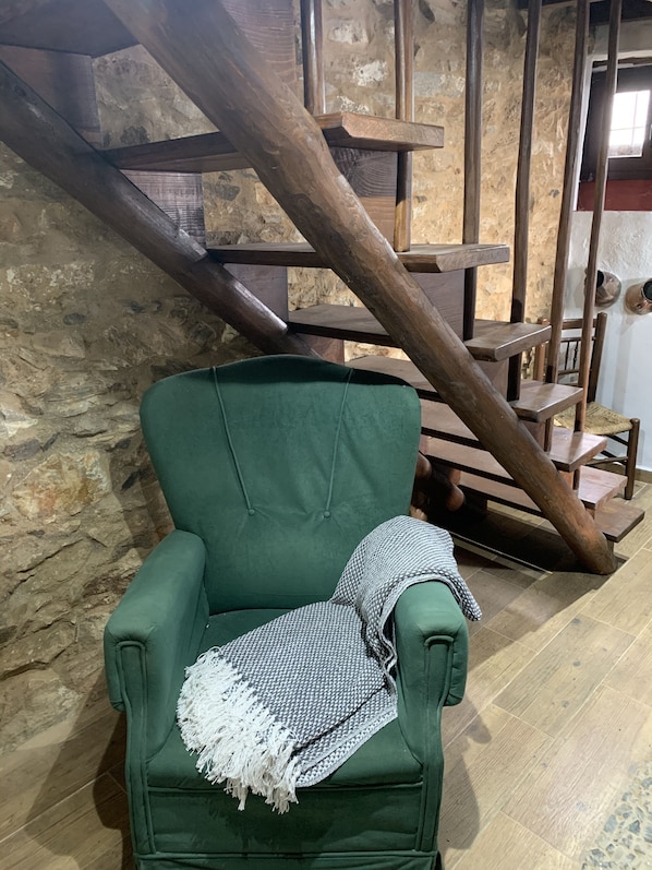 A green armchair with a patterned throw blanket, positioned in front of a rustic stone wall and wooden staircase.