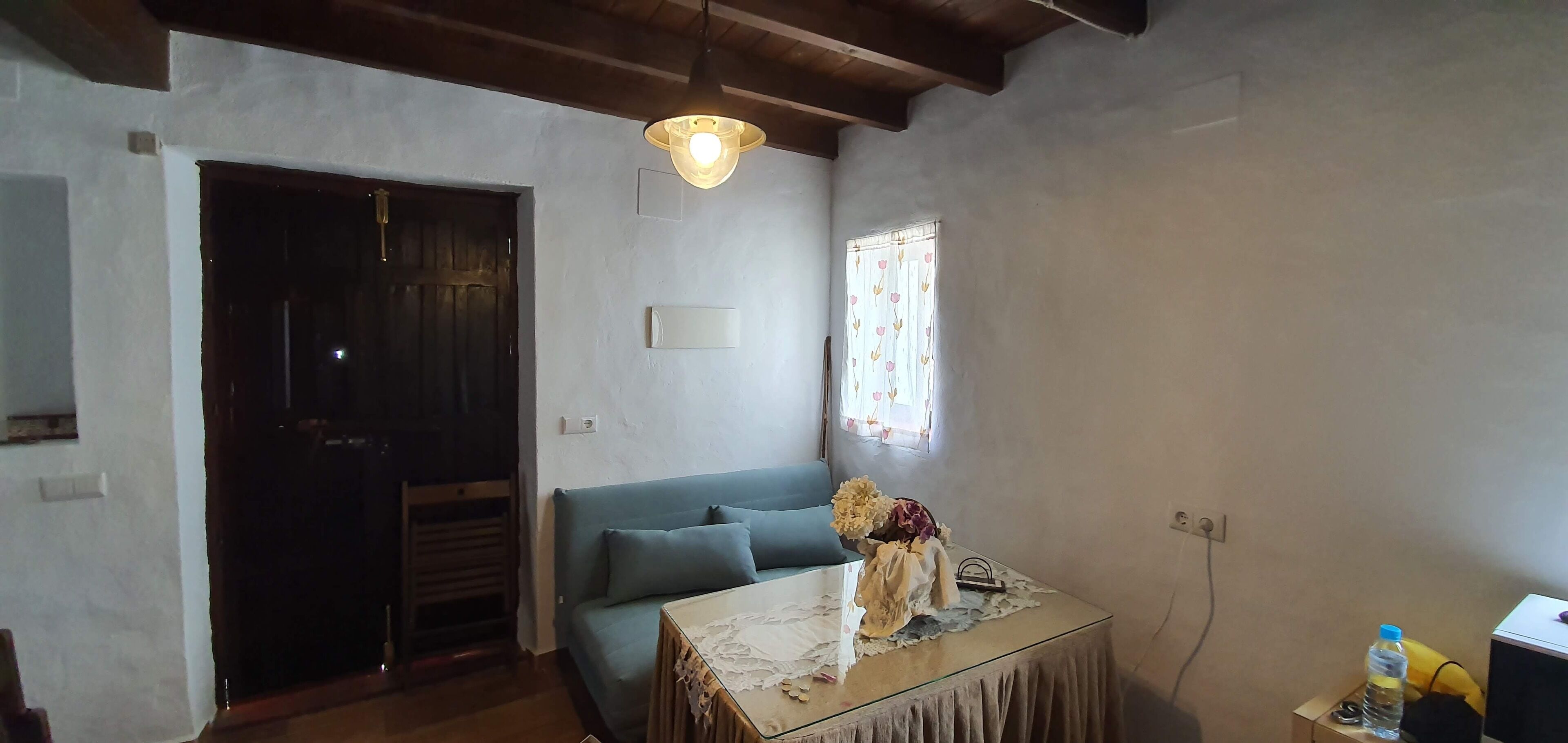 A room with a wooden ceiling, a blue sofa, a glass table with a lace tablecloth, and a window with floral curtains.