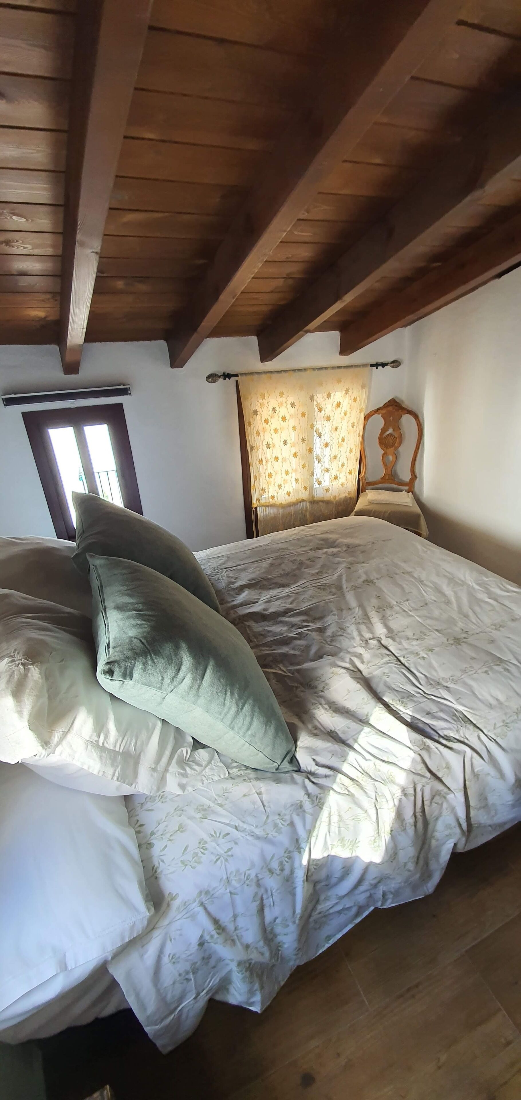 Une chambre avec un plafond en bois, un lit recouvert d’une literie à motifs floraux et une fenêtre avec des rideaux.