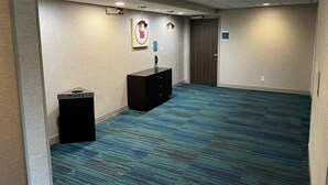 A hotel room with a large carpeted area, a desk with a trash bin, a coat rack, and a door. - Home2 Suites BY Hilton Hinesville (HINESVILLE)