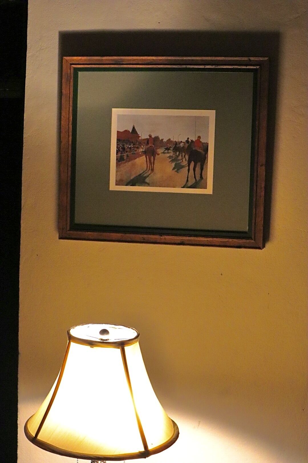 A framed painting of a street scene with people and horses, and a table lamp below it.