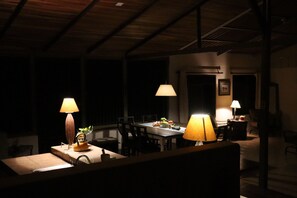 Uma sala de jantar pouco iluminada com duas mesas, cada uma com um abajur. Há um banco em primeiro plano, e o espaço possui um teto alto com vigas aparentes.