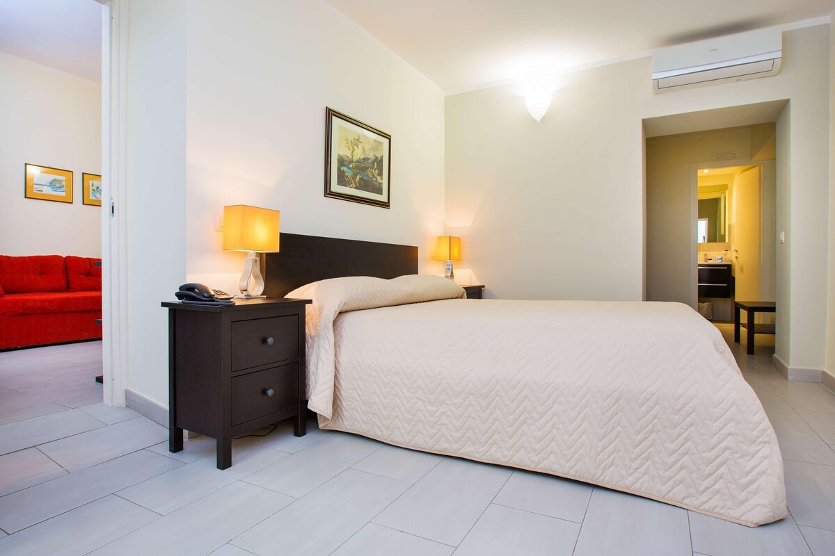 A hotel room with a bed, bedside tables, a red sofa, and a bathroom visible through an open door.