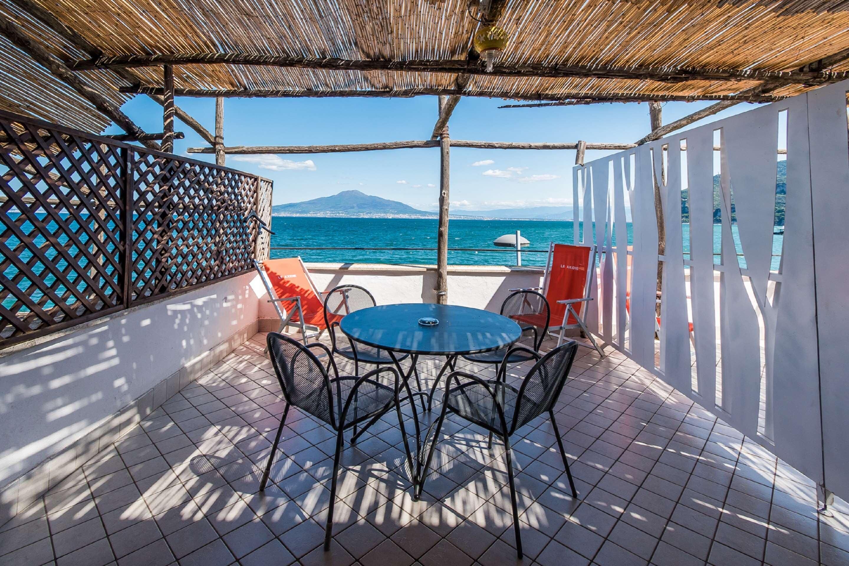 An outdoor patio with a round table and chairs, a thatched roof, and a view of the sea and a mountain.
