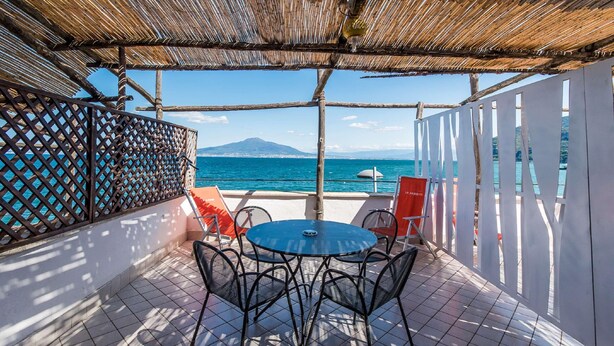 An outdoor patio with a round table and chairs, a thatched roof, and a view of the sea and a mountain. - Residence Le Axidie Sure Hotel Collection by BW (Vico Equense)
