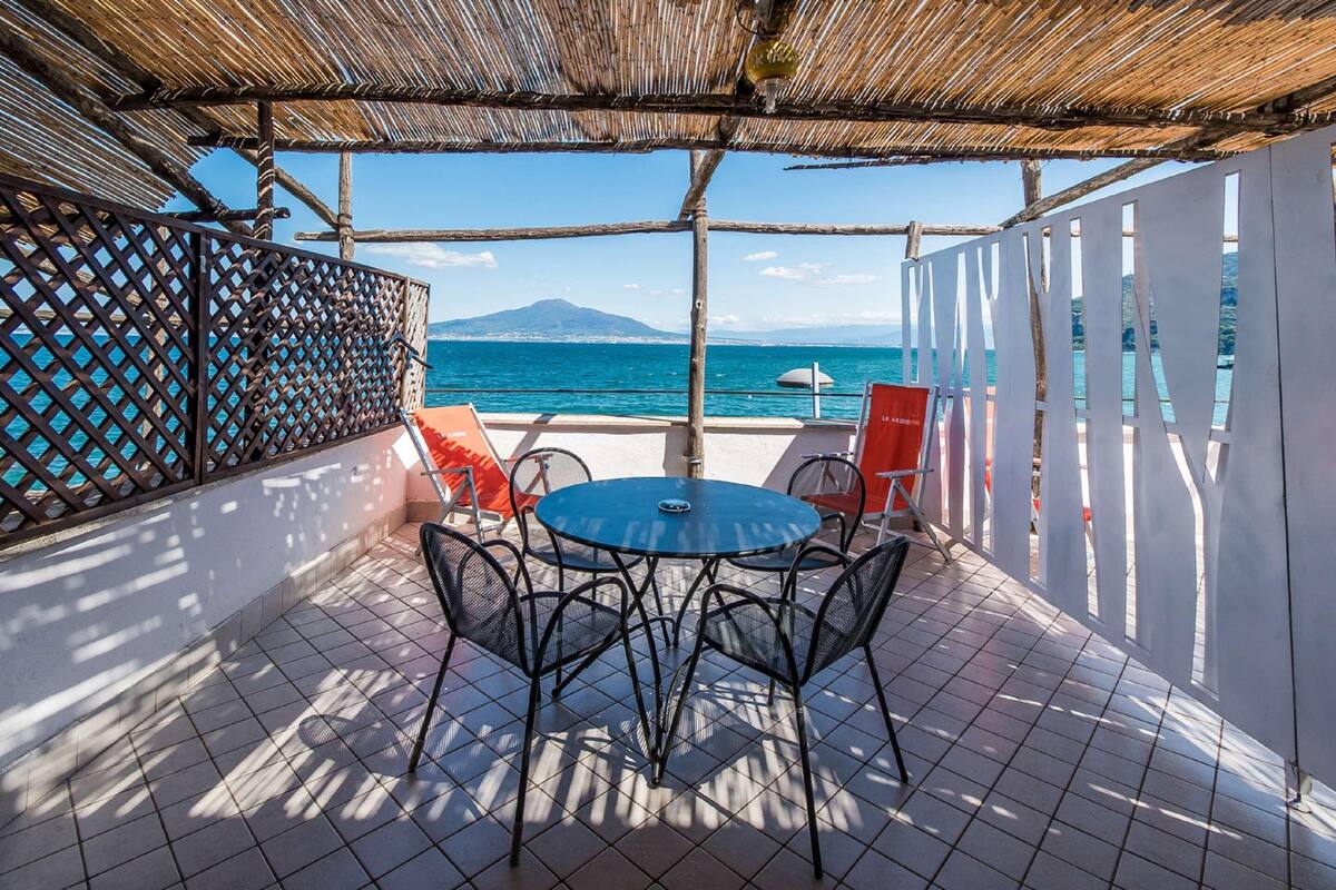 An outdoor patio with a round table and chairs, a thatched roof, and a view of the sea and a mountain.