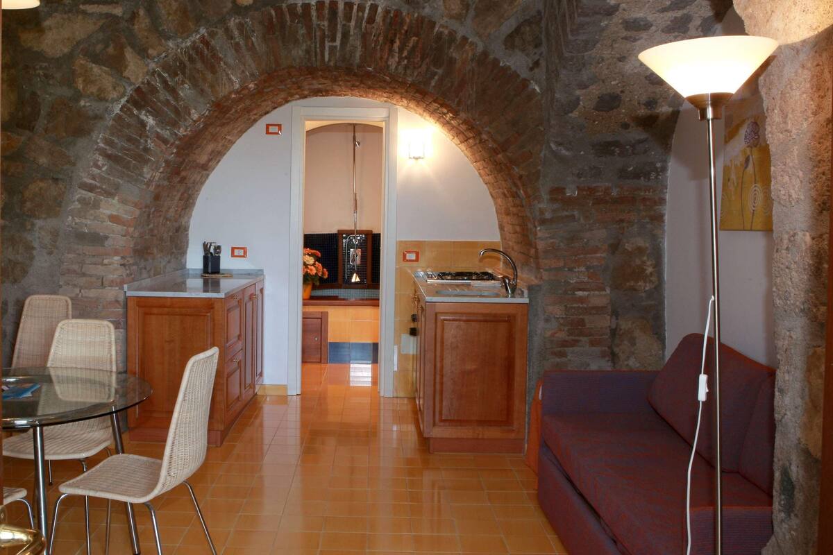 A living room with a stone archway, a dining area with wicker chairs, a sofa, and a kitchen in the background.