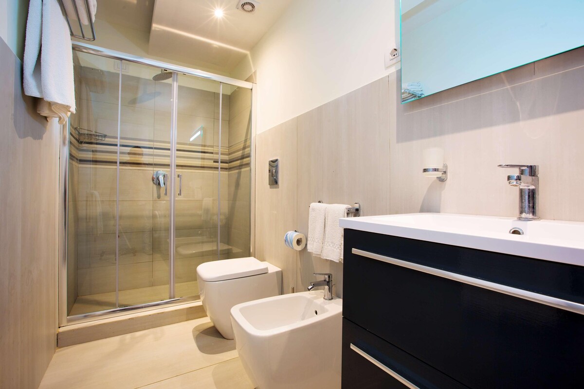 A modern bathroom with a glass shower enclosure, a white toilet, a bidet, and a vanity with a mirror.