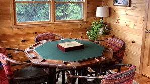 A wooden cabin interior with a round table, four chairs, a window with a view of trees, a ceiling fan, and a framed picture on the wall.