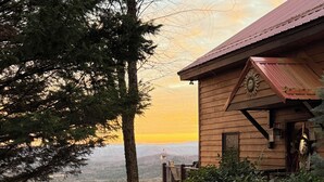 A wooden cabin with a balcony overlooking a forested valley, a gravel path leading to the entrance, and a sign in a foreign language.
