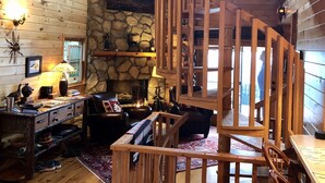A cozy cabin interior with a stone fireplace, wooden walls, a spiral staircase, and a rustic decor.