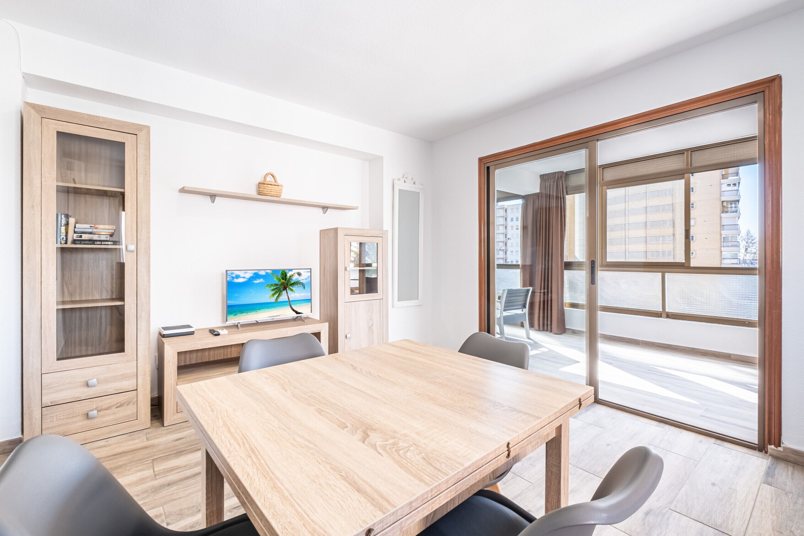 Una habitación moderna con una mesa de comedor de madera, sillas, un televisor, una estantería y un ventanal con vistas a edificios.