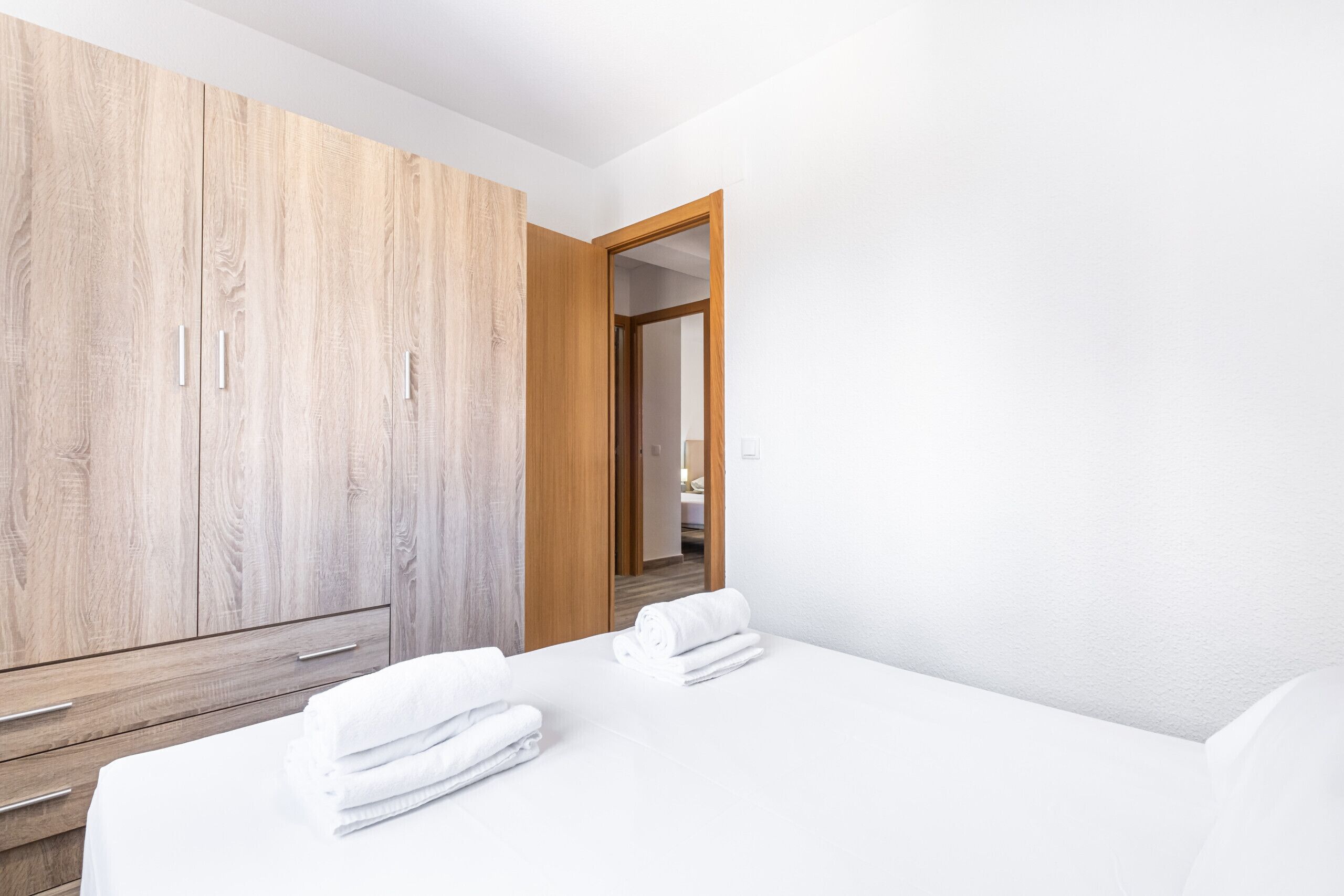 A hotel room with a wooden wardrobe, a bed with white linens, and a view into the adjacent room.
