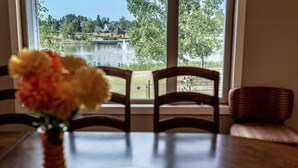 Une table à manger avec des chaises, un vase rempli de fleurs et une vue sur un lac et des maisons, visible à travers de grandes fenêtres.