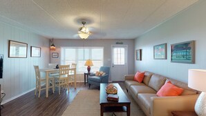 A living room with a dining table, chairs, a sofa, and a coffee table. There are two windows with blinds, a door, and a ceiling fan. - Walk to Sandfest, Shared Pool, Boardwalk to the Beach (Port Aransas)