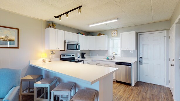 A modern kitchen with white cabinets, a central island, a microwave, an oven, and a dishwasher. There are three stools at the island and a painting on the wall. - Walk to Sandfest, Shared Pool, Boardwalk to the Beach (Port Aransas)