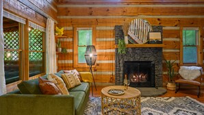 Ein gemütliches Blockhaus-Interieur mit einem grünen Sofa, einem runden Korbtisch, einem Kamin aus Stein und einer Treppe mit Holzwand.