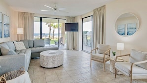 A spacious living room with a large sofa, a round ottoman, and a chair. There is a television mounted on the wall, and a ceiling fan is visible. - Summer House 106B | Ground floor, wrap around deck, direct beach front! (Orange Beach)