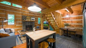 Interior de una cabaña de troncos con chimenea de piedra, escalera de madera y comedor con taburetes.