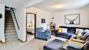 Ein Wohnzimmer mit einer Couch, einem Sessel und einem Hocker. Eine Treppe führt in einen anderen Raum.