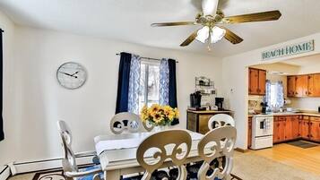 A dining area with a table, chairs, a ceiling fan, a clock, and a kitchen in the background.